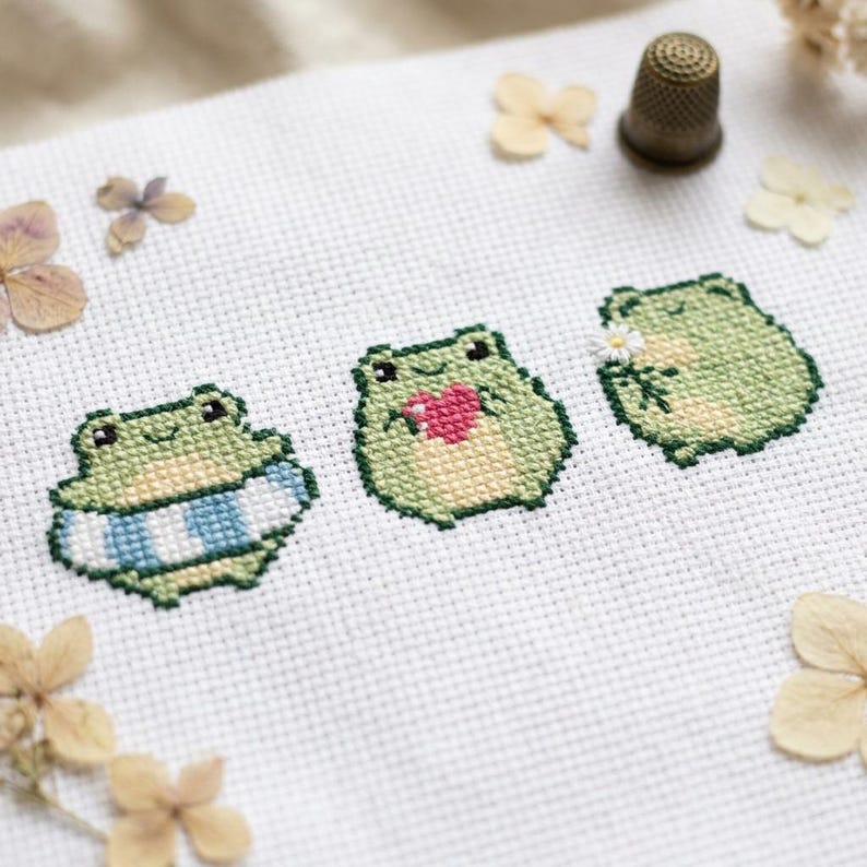 May include: A white Aida fabric featuring three cross-stitched green frogs. The first frog wears a blue and white striped inner tube, the second holds a pink heart, and the third holds a small white flower. A metal thimble sits nearby.