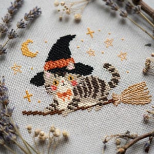Puede incluir: Un bordado de punto de cruz sobre tela gris claro que muestra un gato atigrado con un sombrero de bruja negro con una banda naranja. El gato tiene ojos amarillos, mejillas rosadas y una pajarita naranja, montado en una escoba marrón.