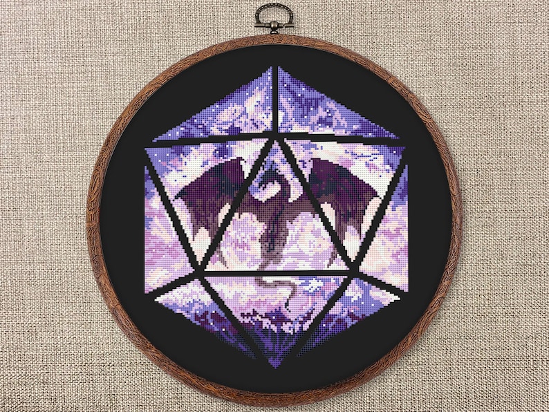 May include: A cross-stitch artwork of a dragon inside a geometric shape, mounted in a wooden embroidery hoop. The dragon is dark purple with outstretched wings, set against a background of light purple and white. The hoop is approximately 10 inches in diameter.