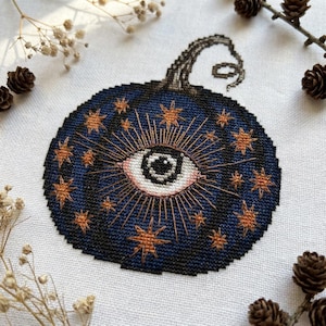 以下が含まれることがあります： 白い布に施されたクロスステッチ刺繍で、濃い青色のカボチャの形をしています。カボチャはオレンジ色の星と、放射状の線を持つ中央の目のようなモチーフで装飾されています。上部からは、巻きひげのある濃い茶色の茎が伸びています。