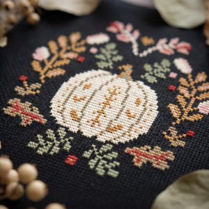 May include: A cross-stitch embroidery on black fabric featuring a central white pumpkin surrounded by a wreath of botanical elements including leaves, sprigs, and small berries in shades of muted green, tan, pink, and deep red.