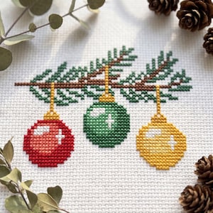 May include: A cross-stitch embroidery on white fabric featuring a brown pine branch with three hanging ornaments in red, green, and yellow. Each round ornament has a gold cap and white highlights. The design is surrounded by pine cones.