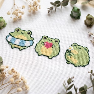 May include: A white fabric piece featuring three cross-stitched green frogs. The first frog wears a blue and white striped inner tube, the second holds a pink heart, and the third holds a small white flower with green leaves and a yellow center.