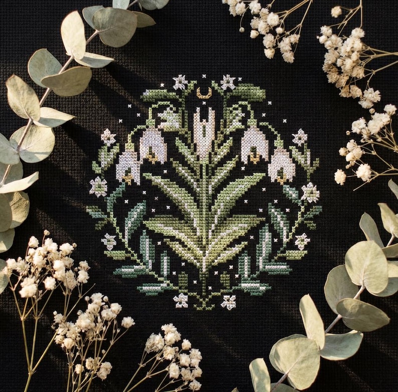 May include: A cross-stitch floral design with white bell-shaped flowers, green leaves, and a crescent moon. The embroidery is on a black background, framed by eucalyptus and white flowers. The design is a circular pattern.