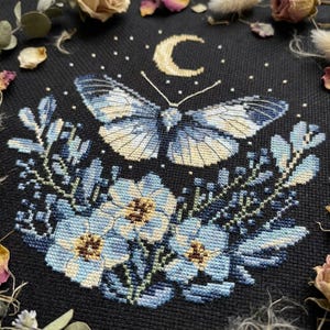 Könnte beinhalten: Kreuzstichmuster mit einem Schmetterling mit blau-cremefarbenen Flügeln, über blauen Blumen und Blättern. Ein Halbmond und Sterne befinden sich über dem Schmetterling, alles auf schwarzem Hintergrund. Das Design ist von getrockneten Blumen umgeben.