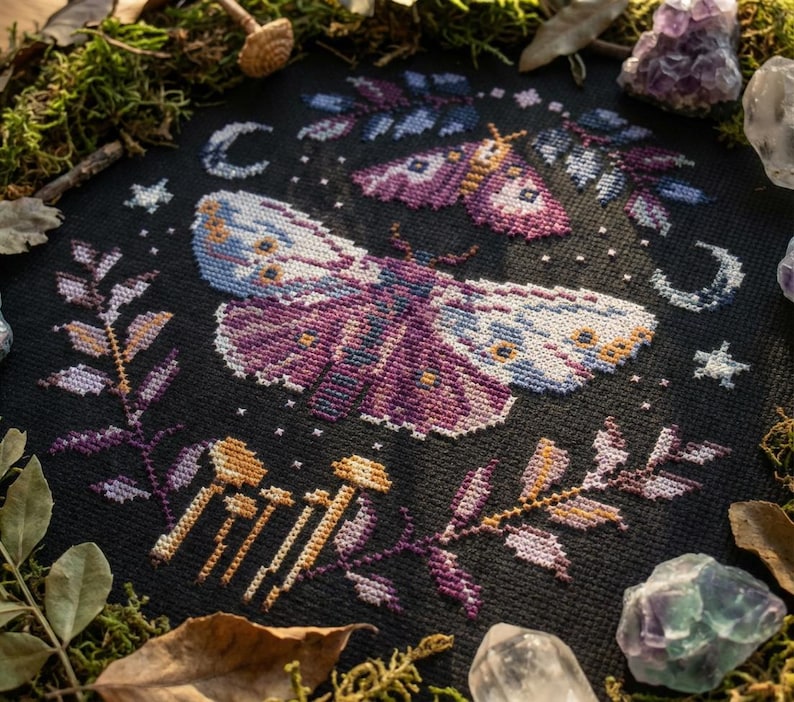 Puede incluir: Dise&ntilde;o de punto de cruz con dos polillas con intrincados patrones de alas en tonos morados, azules y blancos, sobre un fondo negro. El dise&ntilde;o incluye elementos celestiales como lunas y estrellas, con acentos bot&aacute;nicos.