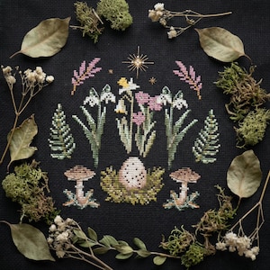Puede incluir: Un dise&ntilde;o de punto de cruz con un arreglo floral central con flores blancas, rosas y amarillas, rodeado de follaje verde y setas. El dise&ntilde;o est&aacute; sobre un fondo negro, enmarcado con hojas secas y musgo.