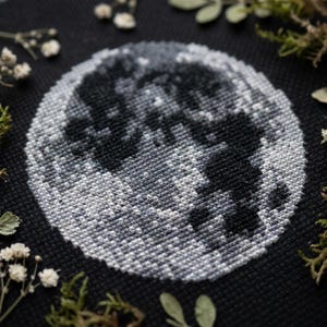 Könnte beinhalten: Ein kreisförmiges Kreuzstich-Stickbild auf schwarzem Stoff, das einen detaillierten Mond in Weiß-, Grau- und Schwarztönen darstellt. Das Design zeigt texturierte Stiche, die die Krater und Oberflächenvariationen der Mondlandschaft nachahmen.