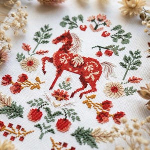 Può includere: Un pezzo di tessuto bianco con un ricamo a punto croce di un cavallo rosso circondato da elementi botanici tra cui foglie verdi, bacche rosse e fiori beige. Il disegno è centrato sul tessuto con composizioni floreali simmetriche.
