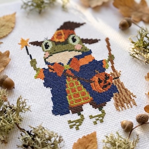 May include: A cross-stitch embroidery on white fabric featuring a green frog dressed as a wizard. The frog wears a brown hat, a blue robe with an orange bow tie, and a plaid vest. It holds a yellow star wand and a brown broom with a pumpkin.
