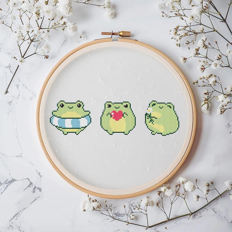 May include: A circular wooden embroidery hoop containing white fabric with a cross-stitch pattern of three light green frogs. The first frog wears a blue and white striped inner tube, the second holds a red heart, and the third holds a white flower.