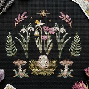 Puede incluir: Un dise&ntilde;o de punto de cruz con un huevo en un nido en el centro, rodeado de flores, setas y follaje. La paleta de colores incluye rosa, blanco, verde y marr&oacute;n sobre un fondo negro. Tambi&eacute;n hay una estrella dorada y ramitas de lavanda.