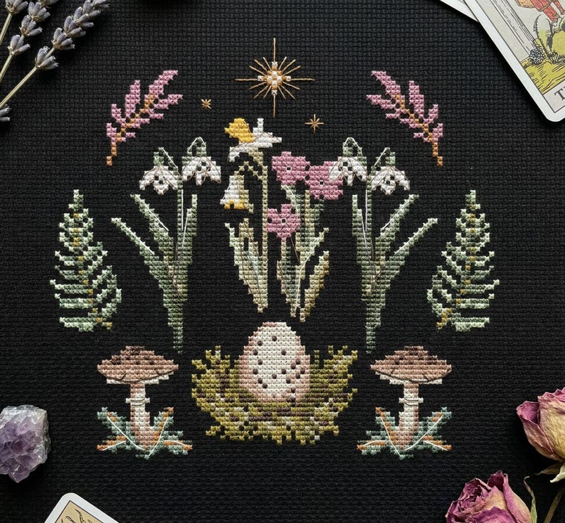 Puede incluir: Un dise&ntilde;o de punto de cruz con un huevo en un nido en el centro, rodeado de flores, setas y follaje. La paleta de colores incluye rosa, blanco, verde y marr&oacute;n sobre un fondo negro. Tambi&eacute;n hay una estrella dorada y ramitas de lavanda.