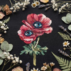 May include: Cross-stitch artwork of two red anemone flowers with blue eye-like centers, set against a black background. The flowers are surrounded by dried flowers and greenery. The piece has a handmade, botanical theme.