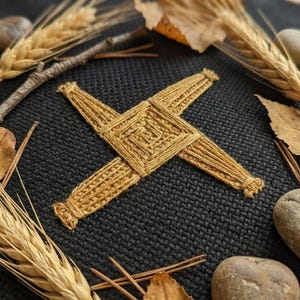 Puede incluir: Una cruz de Santa Brígida de paja tejida, un símbolo irlandés tradicional, está centrada en una superficie texturizada de color gris oscuro. La cruz está rodeada de espigas de trigo, hojas y pequeñas piedras, creando una estética rústica y natural.