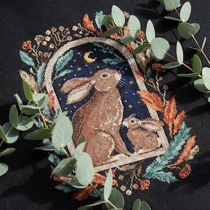 Könnte beinhalten: Ein Kreuzstich-Design mit zwei braunen Hasen in einem bogenförmigen Rahmen vor einem sternenklaren Nachthimmel. Der Rahmen ist von farbenfrohem Laub in Orange-, Grün- und Beigetönen umgeben, auf schwarzem Hintergrund.