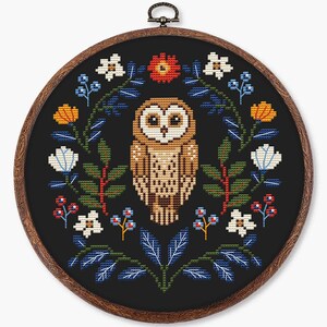 May include: Cross-stitch embroidery of an owl surrounded by a floral wreath. The owl is brown and tan, with large eyes. The wreath includes white, orange, and blue flowers, and green leaves, all on a black background, in a wooden hoop.