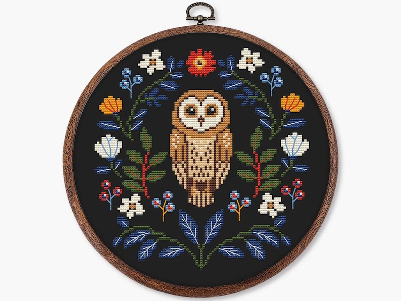 May include: Cross-stitch embroidery of an owl surrounded by a floral wreath. The owl is brown and tan, with large eyes. The wreath includes white, orange, and blue flowers, and green leaves, all on a black background, in a wooden hoop.