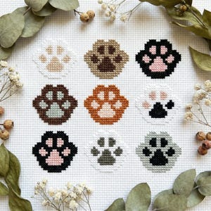May include: Nine cross-stitch paw prints in various colours, including white, brown, black, and grey, are arranged on a white fabric background. The paw prints are surrounded by dried leaves and small white flowers, creating a craft-themed design.