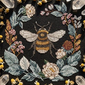 May include: Cross-stitch embroidery of a bee with a floral wreath on a black background. The bee is detailed with yellow and brown stripes and white wings. The wreath includes white roses, green leaves, and small pink flowers. Crystals and dried flowers surround the image.