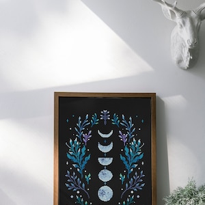 May include: A framed cross-stitch embroidery featuring a black background with a floral wreath design. The wreath is made up of green and purple leaves and flowers. In the center of the wreath are five blue and white moons in different phases.