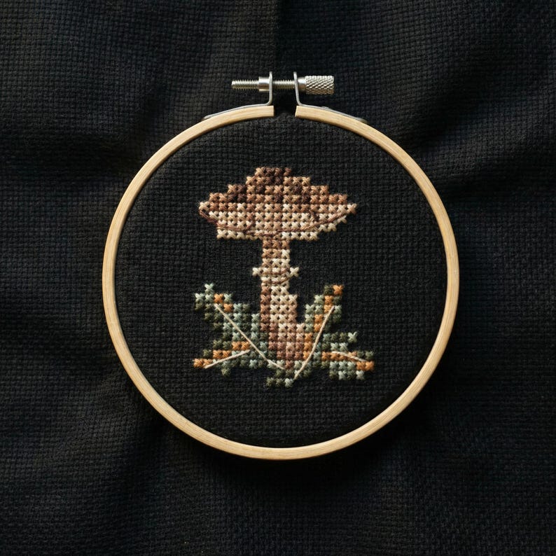 Puede incluir: Bordado de punto de cruz de un hongo en un aro de madera. El hongo es marr&oacute;n y beige, con follaje verde y beige. El fondo es negro.