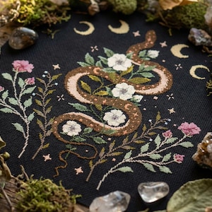 Puede incluir: Un diseño de punto de cruz sobre tela negra presenta una serpiente marrón y bronceada entrelazada con flores blancas y flores rosas. Crecientes lunares y estrellas están dispersos por el fondo. El diseño está rodeado de musgo, cristales y follaje.