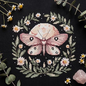 May include: Cross-stitch design featuring a moth with pink and cream wings, centered on a cream-colored moon. The moth is surrounded by a wreath of green leaves and white daisies, set against a black background. A pink crystal and sprigs of eucalyptus and daisies are in the corners.