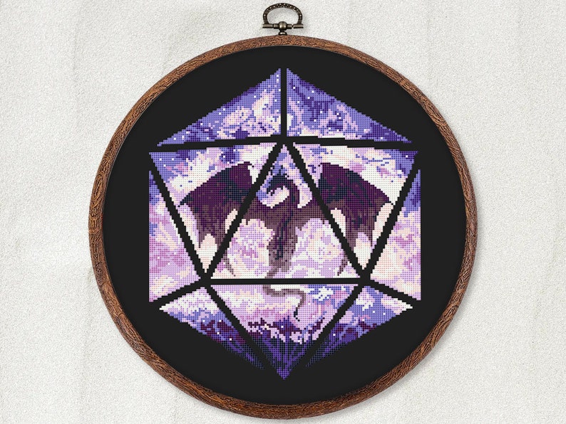 May include: A cross-stitch design of a dragon inside a D20 die, framed in a wooden hoop. The dragon is dark purple, set against a light purple and white background. The die is outlined in black.