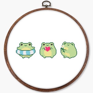 May include: A circular wooden embroidery hoop containing a white fabric panel with a cross-stitch pattern of three light green frogs. The first frog wears a blue and white striped inner tube, the second holds a pink heart, and the third holds a flower.