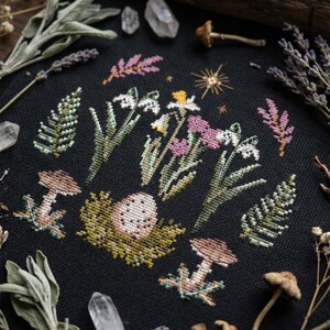 Puede incluir: Bordado de punto de cruz sobre tela negra con una escena del bosque con setas, flores y un huevo en un nido. El dise&ntilde;o incluye flores blancas, rosas y amarillas, helechos verdes y setas marrones. Cristales y plantas secas rodean el bordado.