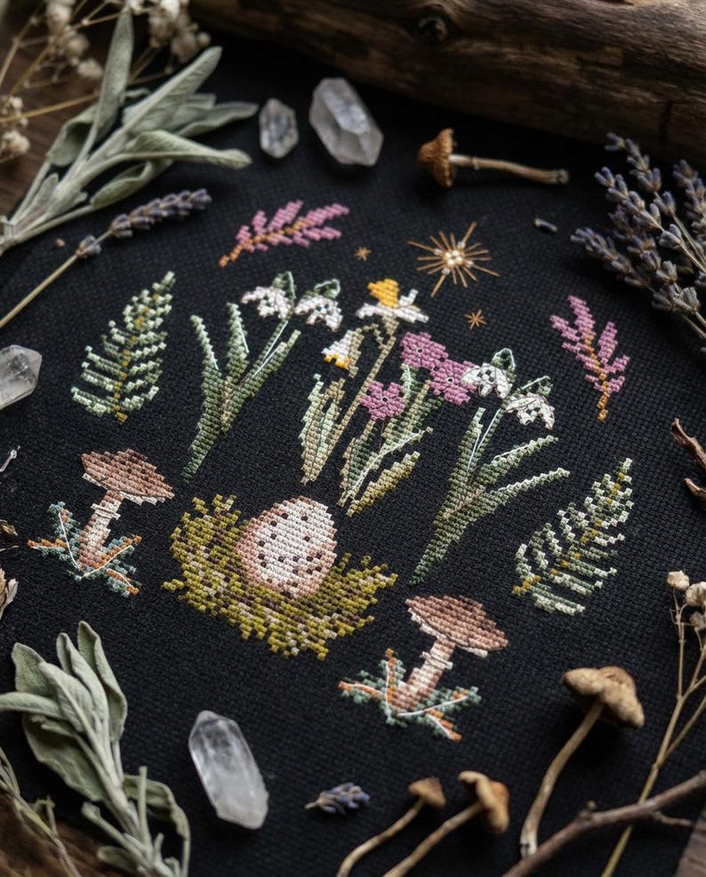 Puede incluir: Bordado de punto de cruz sobre tela negra con una escena del bosque con setas, flores y un huevo en un nido. El dise&ntilde;o incluye flores blancas, rosas y amarillas, helechos verdes y setas marrones. Cristales y plantas secas rodean el bordado.