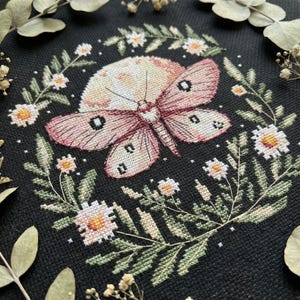 May include: Cross-stitch design featuring a moth with pink and cream wings, set against a full moon and surrounded by floral elements. The design is stitched on black fabric, with details in white, green, and orange.