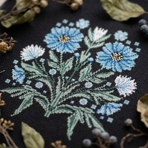 Könnte beinhalten: Ein Kreuzstich-Blumendesign mit blauen Blumen mit gelben Zentren und weißen Akzenten auf schwarzem Hintergrund. Die Blätter sind grün und das Design ist von getrocknetem Laub umgeben.