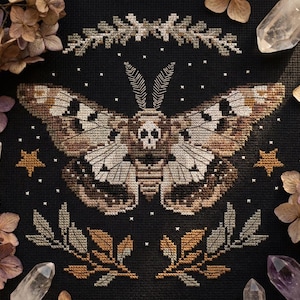 May include: Cross-stitch design featuring a large moth with a skull on its body, set against a black background. The moth's wings are detailed with brown, tan, and white patterns. The design includes stars, crystals, and floral accents.