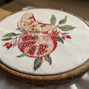 May include: An embroidered artwork in a wooden hoop, depicting a moth and pomegranate. The moth has tan and brown wings, while the pomegranate is red and cream. Green leaves and red berries surround the fruit. The hoop is approximately 20 cm in diameter.