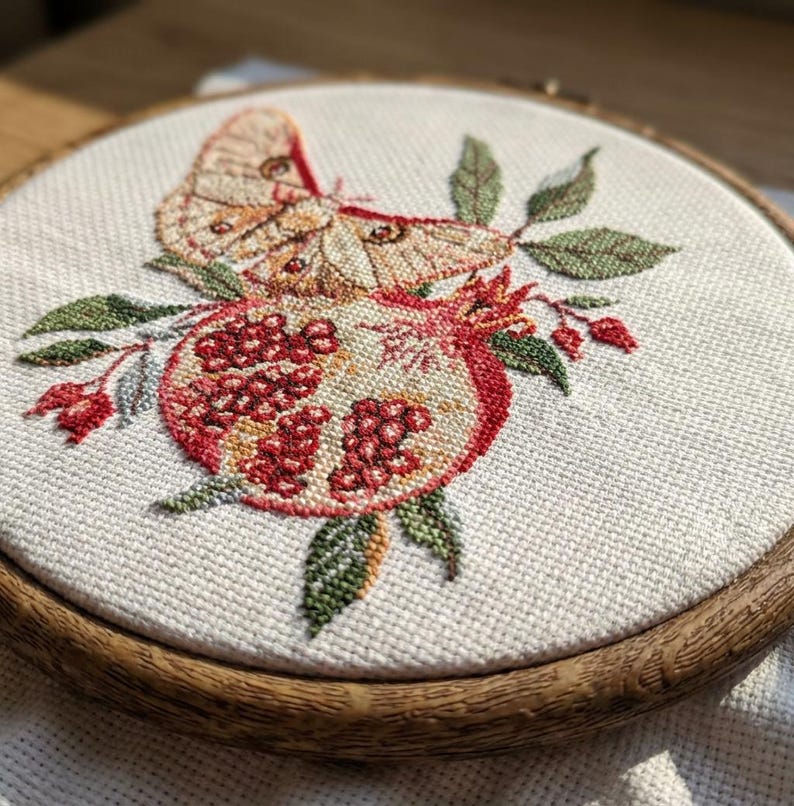 May include: An embroidered artwork in a wooden hoop, depicting a moth and pomegranate. The moth has tan and brown wings, while the pomegranate is red and cream. Green leaves and red berries surround the fruit. The hoop is approximately 20 cm in diameter.