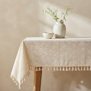 May include: A beige rectangular tablecloth featuring a repeating white daisy floral pattern and vertical stripes. The edges are finished with a decorative fringe of small, hanging tassels. A ceramic vase with white flowers sits on top.
