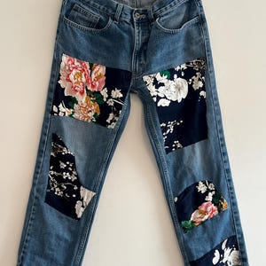 May include: Blue denim jeans with floral patches on the front. The patches are in various sizes and shapes and feature a variety of floral patterns.
