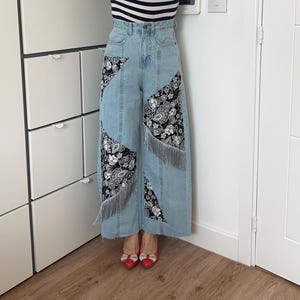 Può includere: Pantaloni in denim blu chiaro con pannelli in tessuto a fantasia paisley bianco e nero e bordi con frange.