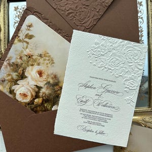 Könnte beinhalten: Elegante Hochzeitseinladungssuite mit einer cremefarbenen Einladung mit geprägten floralen Details und Schrifttext. Ein brauner Umschlag mit passenden geprägten Rosen enthält einen floral bedruckten Einsatz. Die Einladung nennt Stephen Guerrero und Emily Welbourn.