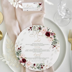 May include: A round white menu card with a floral design featuring burgundy and pink flowers. The menu card has a gold border and the text "Menu" in gold lettering. The menu includes salad, mains, and dessert options.