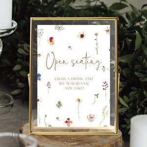 May include: A gold-framed sign with a white background and floral illustrations. The sign reads "Open seating. Grab a drink and sit wherever you like!"