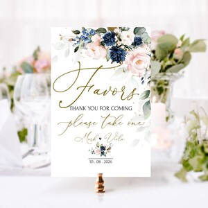 May include: A white sign with a floral design in shades of blue and pink. The sign says "Favors" in gold script and "Thank you for coming, please take one, Mark & Vila, 10.08.2026".