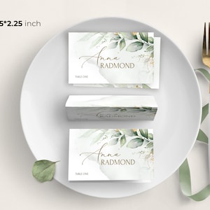 May include: A white place card with a green and gold floral design. The text "Anna Radmond" is written in gold script. The place card is 3.75 inches by 2.25 inches and says "Table One" at the bottom.