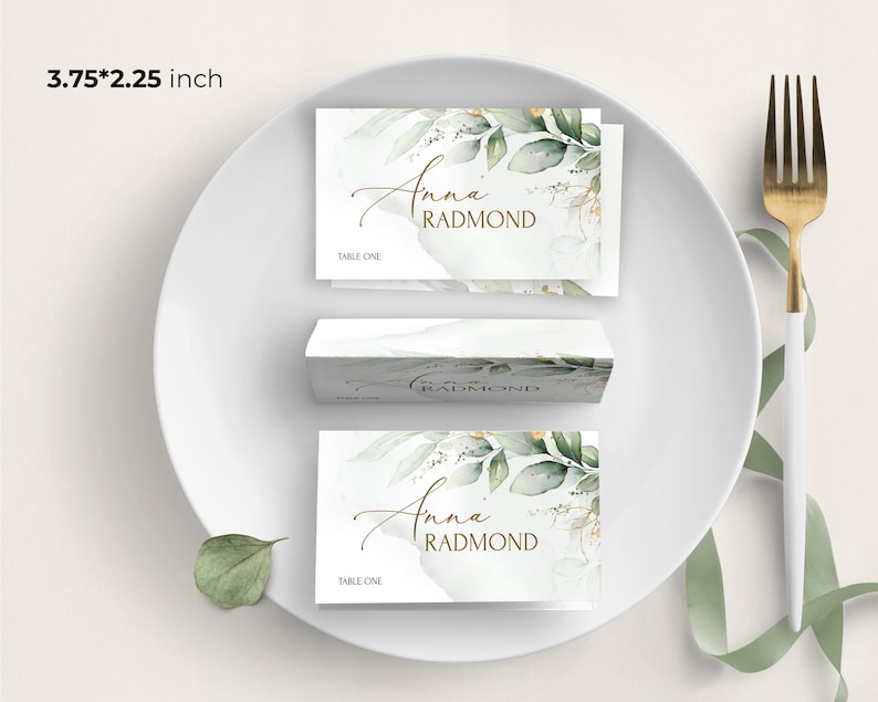 May include: A white place card with a green and gold floral design. The text "Anna Radmond" is written in gold script. The place card is 3.75 inches by 2.25 inches and says "Table One" at the bottom.
