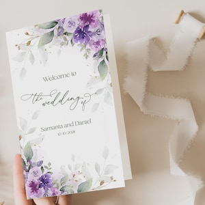 May include: A wedding program with a floral watercolour design in purple and green. The text reads "Welcome to the wedding of Samanta and Daniel 10/10/2028". A white ribbon is next to the program.
