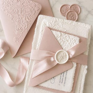 May include: A set of stationery featuring a dusty rose envelope with a floral embossed flap, a white deckle-edge card, and a dusty rose card secured with a dusty rose satin ribbon and a white wax seal featuring a floral design.