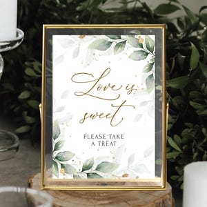 Peut inclure: Une pancarte encadrée d'or avec un fond blanc et des feuilles vertes en aquarelle. Le texte sur la pancarte dit "L'amour est doux. Prenez un régal."