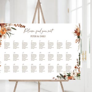 May include: A large white seating chart with the text "Please, find your seat" and the names "PETER & EMILY." The chart is decorated with floral arrangements in shades of orange, brown, and white. The chart is displayed on a wooden easel.